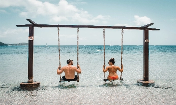 Una pareja de espaldas, mirándose, sentados en un columpio en la orilla de una playa paradisíaca, de aguas transparentes