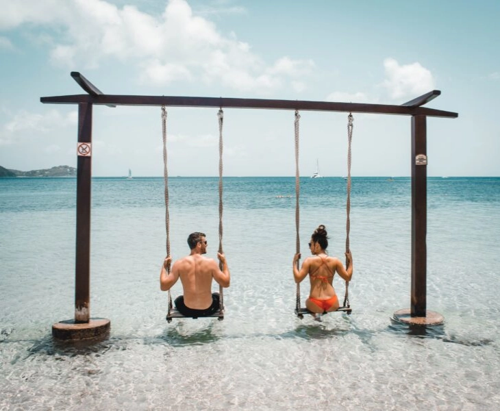 Una pareja de espaldas, mirándose, sentados en un columpio en la orilla de una playa paradisíaca, de aguas transparentes