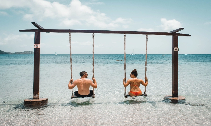 Una pareja de espaldas, mirándose, sentados en un columpio en la orilla de una playa paradisíaca, de aguas transparentes