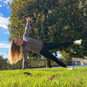 Una chica en un parque, sobre el césped, haciendo una plancha lateral, con un brazo y una pierna estiradas hacia el cielo.