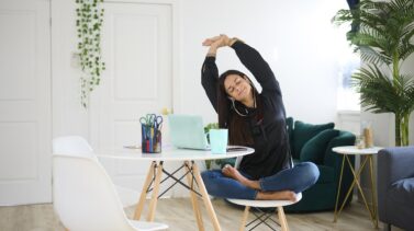 Smiling Latina businesswoman sitting at desk stretching. Estiramientos para los que pasan muchas horas sentados.
