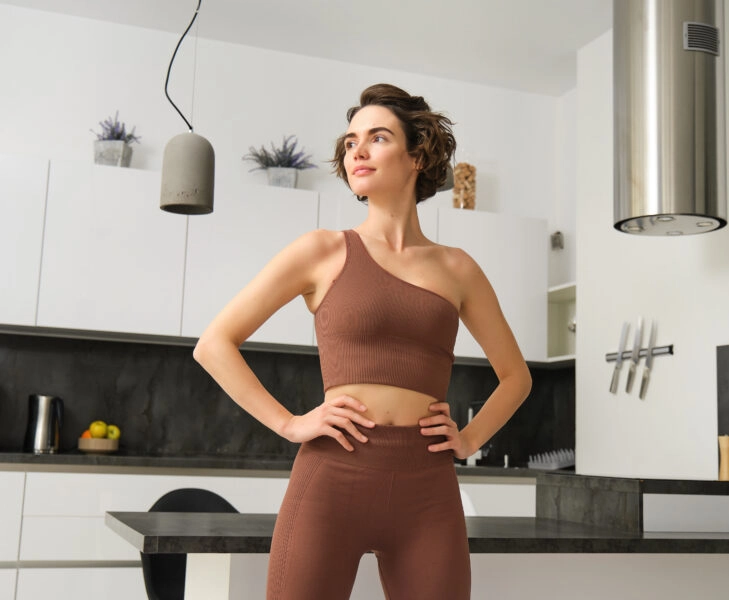 Image of young fit woman, fitness girl in sportswear, standing in power pose, workout at home, looking outside window, training alone indoors