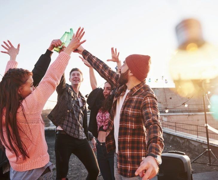 Decorative festive light bulbs. Holidays on the rooftop. Cheerful group of friends raised their hands up with alcohol