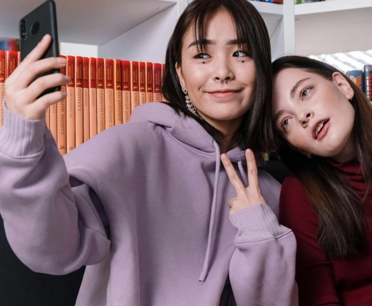 Dos chicas haciéndose un selfie; una sonríe y la otra pensando no me reconozco en las fotos, sorprendida al verse distinta
