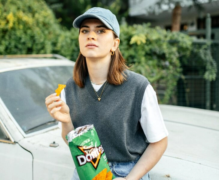 Mujer comiendo snacks frente a un coche, simbolizando cómo los superestímulos de la industria alimentaria se basan en la manipulación para crear adicciones.