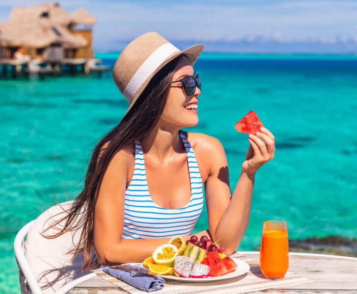 Luxury resort vacation woman eating fruits detox breakfast on beach hotel room with private terrace. Tourist girl healthy diet lifestyle. Frutas tropicales huella de carbono