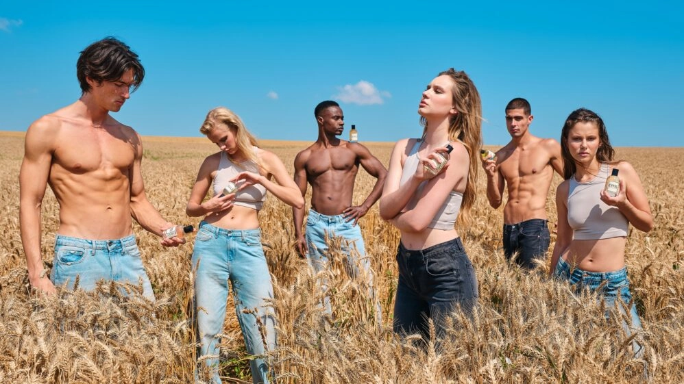 Varios modelos de torso desnudo sostienen frascos de perfume recargables mientras posan entre pacas de paja en un entorno campestre