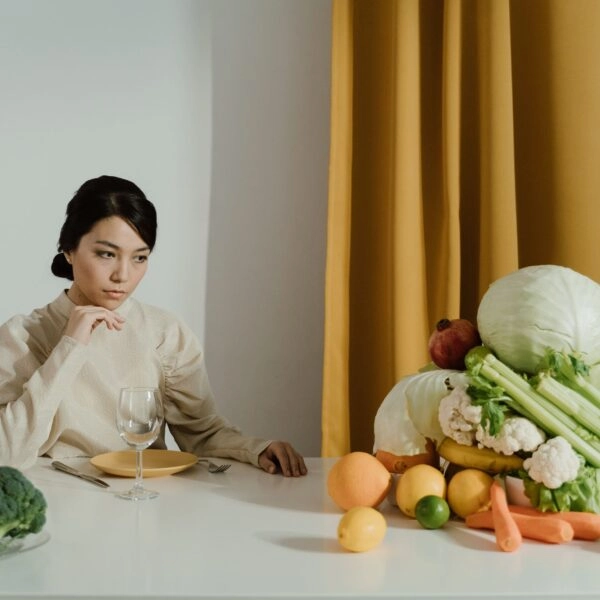 Mujer sentada ante una mesa repleta de frutas y verduras, con gesto pensativo y un plato vacío delante, evocando la presión de tener que comer siempre de forma perfectamente saludable