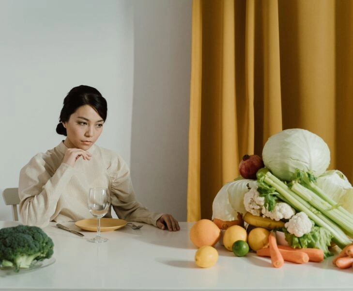 dieta antiinflamatoria que inflama Mujer sentada ante una mesa repleta de frutas y verduras, con gesto pensativo y un plato vacío delante, evocando la presión de tener que comer siempre de forma perfectamente saludable