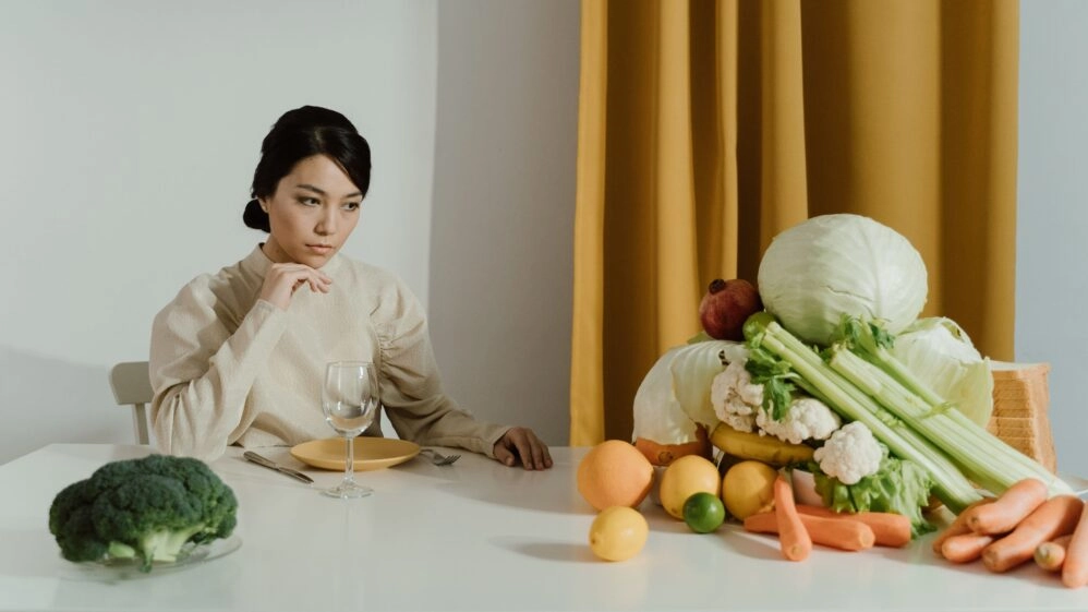 Mujer sentada ante una mesa repleta de frutas y verduras, con gesto pensativo y un plato vacío delante, evocando la presión de tener que comer siempre de forma perfectamente saludable
