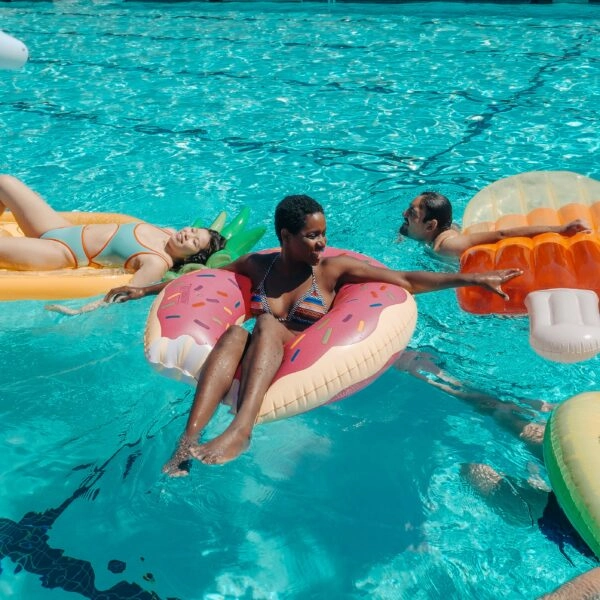 Personas disfrutando de una piscina con flotadores de comida, una metáfora visual de cómo en vacaciones podemos comer peor pero sentirnos menos hinchados