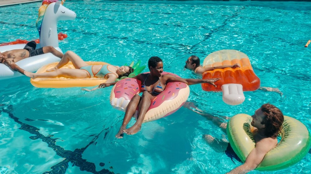 Personas disfrutando de una piscina con flotadores de comida, una metáfora visual de cómo en vacaciones podemos comer peor pero sentirnos menos hinchados
